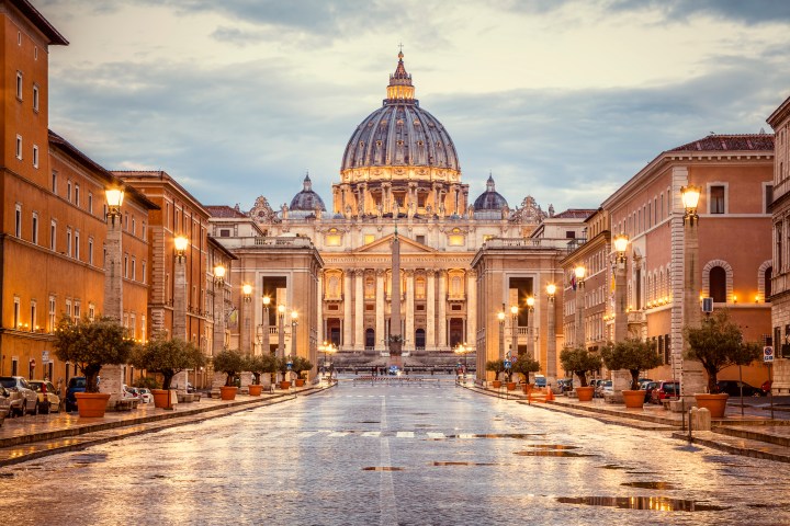 View of St. Peter's Basilica from a wet street lined with lampposts and historic buildings at dusk.