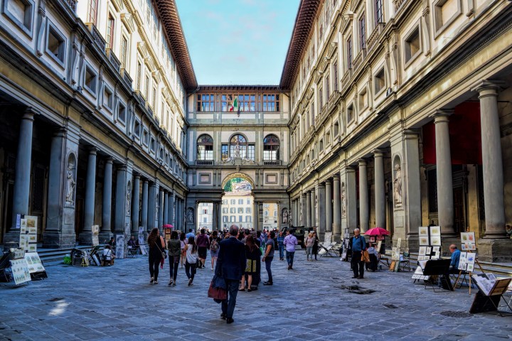 People walking in a narrow street between two tall, historic buildings with art vendors and statues.