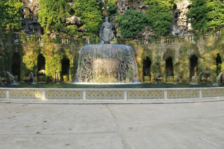 Ornate stone fountain with cascading water, surrounded by greenery and ivy-covered arches.