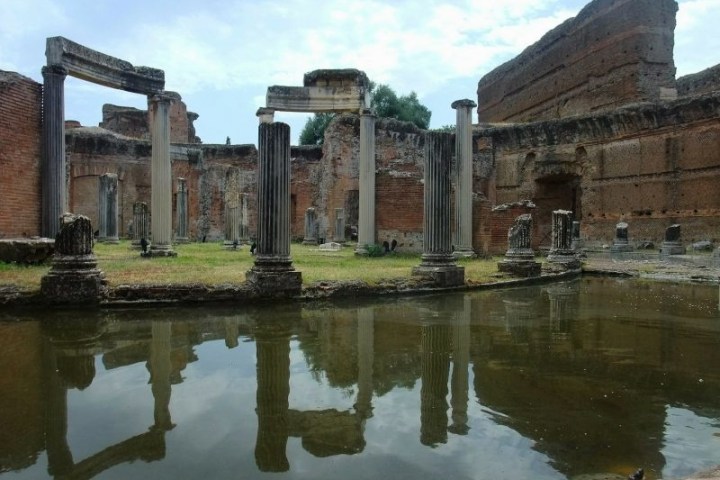 Ancient Roman ruins with columns reflected in a pool of water under a cloudy sky.