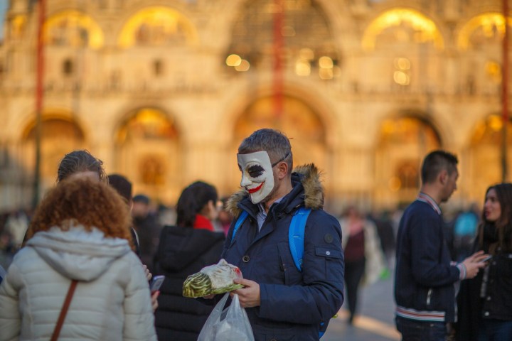Person with painted face holds plastic bag in crowded outdoor setting with blurred building in background.