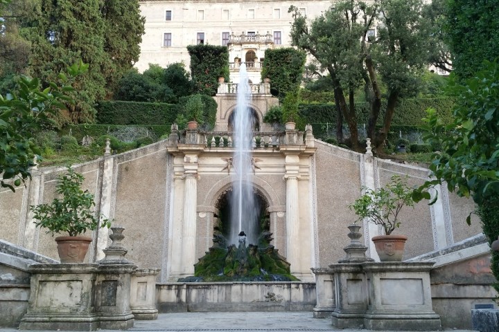 Fountain with water cascading from a stone structure surrounded by greenery and potted plants.